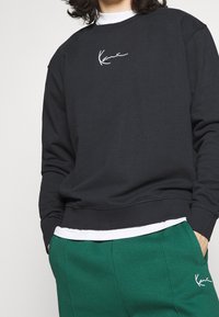 Sudadera negra de algodón con cuello redondo y bordado de logo blanco. Combinada con pantalones de chándal verdes, de corte holgado y con bolsillos.