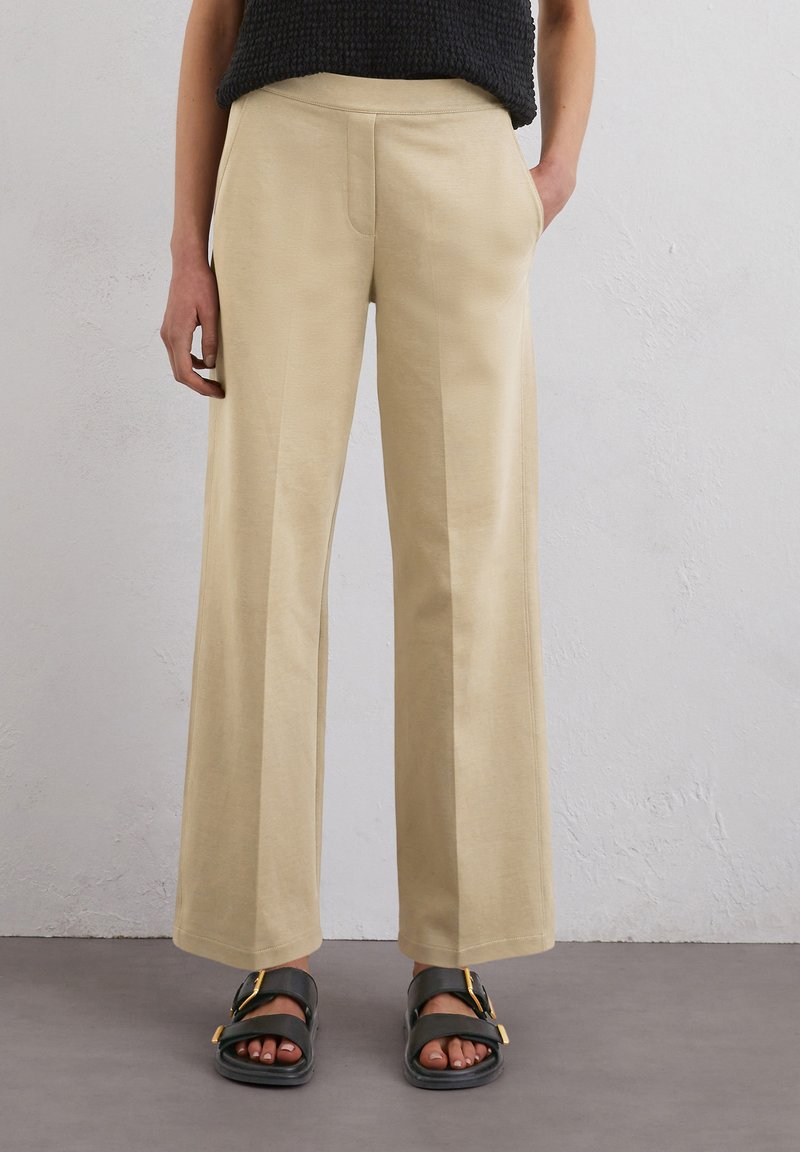 Pantaloni beige a gamba ampia con una texture liscia, due tasche laterali e un design pulito. Abbinati a sandali scuri con un accento dorato.