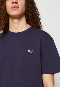 T-shirt in cotone blu navy con un piccolo patch rettangolare ricamato con il testo "TOMMY JEANS", strisce bianche e rosse sopra e sotto.