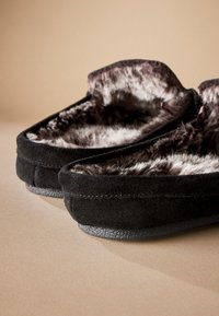 Zapatillas de ante negro con un interior suave forrado de piel. Suelas de goma texturizadas que mejoran el agarre, con detalles de costura a lo largo de los bordes.