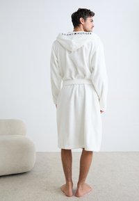 Accappatoio bianco con cappuccio realizzato in tessuto peluche, con cintura annodata e logo sul retro. Superficie piana e texture del pavimento leggera visibile.