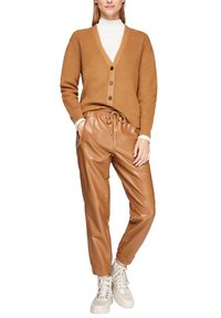 Brauner gerippter Cardigan mit V-Ausschnitt, vier Knöpfen. Braune Jogginghosen aus Kunstleder mit Kordelzug. Hellfarbene Sneakers mit strukturiertem Sohle.