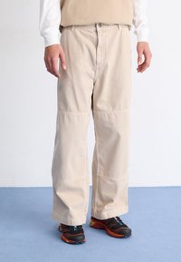 Pantalones de pierna ancha beige con un ajuste relajado, costuras sutiles y cierre de botón. Combinados con zapatillas negras y rojas.