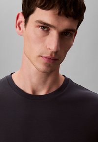 Dunkelgraues, eng anliegendes T-Shirt mit rundem Ausschnitt. Weicher, glatter Stoff. Nahaufnahme des Gesichts eines männlichen Models mit neutralem Gesichtsausdruck und kurzem Haar.