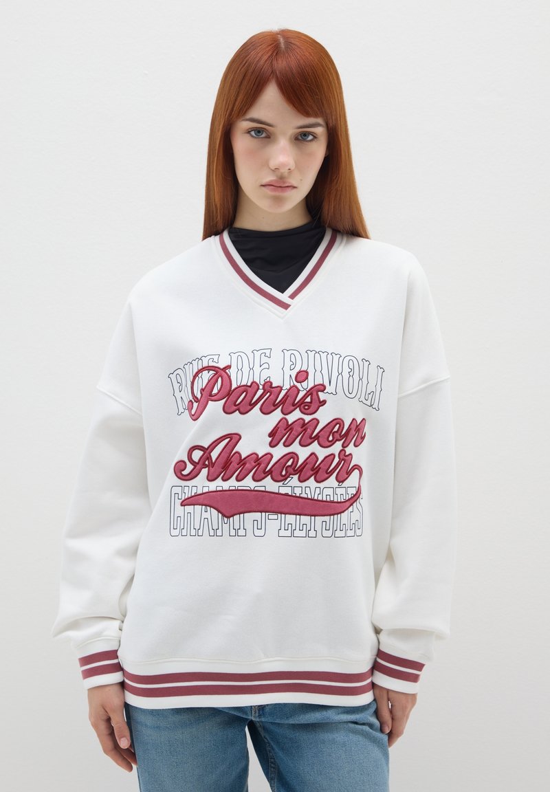 Felpa bianca oversize con scollatura a V, accenti rossi e blu navy, caratterizzata da testo ricamato "Paris mon Amour" e polsini a righe.