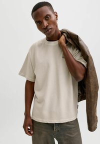 Mladi moški drži rjavo usnjeno jakno čez ramo, nosi bež majico in temne kavbojke, stoji pred enostavnim svetlim ozadjem.