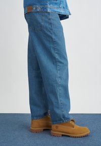 Calças de ganga azul claro com um corte relaxado, combinadas com botas de tornozelo bege com ilhós de metal e uma sola texturizada.