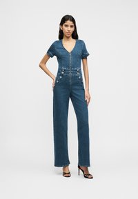 Τζιν ολόσωμη φόρμα με κοντά μανίκια, V-λαιμόκοψη, φερμουάρ μπροστά και μεταλλικά κουμπιά στη μέση· φαρδιά γραμμή παντελονιού σε μεσαίο μπλε χρώμα.