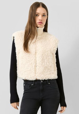 Jeune femme aux cheveux longs porte une veste sherpa sans manches couleur crème par-dessus un haut noir à manches longues et un jean noir, debout devant un fond uni.