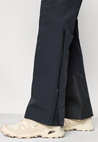 Μαύρο αδιάβροχο παντελόνι με πλαϊνά φερμουάρ, συνδυασμένο με ανοιχτό μπεζ αθλητικά παπούτσια με υφασμένα στοιχεία και στιβαρή σόλα.