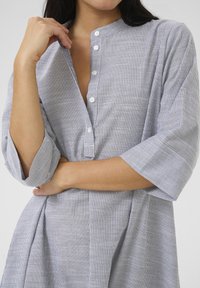 Rochie cu dungi gri și albe, cu guler înălțat, mâneci trei sferturi și un design cu fuste plisate. Textura materialului este netedă.