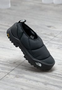 Black slip-on shoe with padded, quilted upper and rubber sole. Features The North Face logo and a rugged tread for grip. Lightweight design.