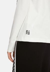 Hvid skjorte med lange ærmer og ribbet struktur samt sort grafisk print ved kanten, kombineret med sorte bukser med geometrisk mønster.