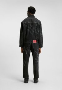 Zwarte spijkerjas en -broek met een geometrisch vervagend patroon op de achterkant. Kenmerkt een rode logo-label aan de taille. Geprofileerde afwerking.