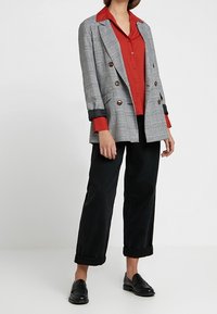 Blazer croisé à carreaux gris, chemise rouge vif en dessous, pantalon noir retroussé aux chevilles, et mocassins noirs. Détails du tissu texturé visibles.