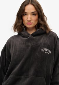 Sudadera de gran tamaño en un gris oscuro de pana, con un bolsillo frontal y el logo bordado de "Superdry" en el pecho. Textura suave y detalles en la capucha.