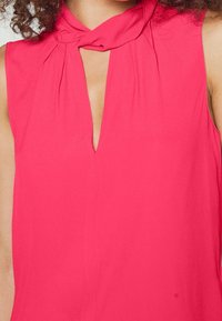 Top senza maniche rosa con scollatura a chiave e dettaglio colletto annodato. Tessuto morbido e liscio con una leggera texture.