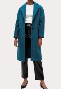 Abrigo de lana color azul verdoso con solapa con muesca, dos bolsillos delanteros y dos botones; combinado con pantalones negros brillantes y tacones negros.