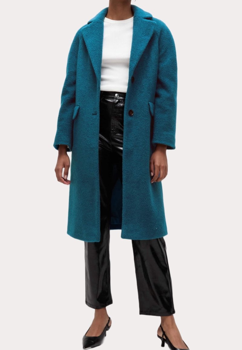 Abrigo de lana color azul verdoso con solapa con muesca, dos bolsillos delanteros y dos botones; combinado con pantalones negros brillantes y tacones negros.