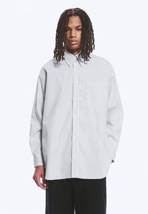 Junger Mann mit langen Dreadlocks, der ein übergroßes weißes Hemd mit dünnen vertikalen grauen Streifen und schwarze Hose vor einem einfachen Hintergrund trägt.