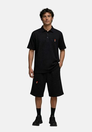 Homme debout portant un polo noir à manches courtes et un short noir jusqu’aux genoux avec des chaussettes noires et des baskets noires.