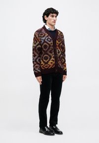 Πλεκτό cardigan σε μπορντό χρώμα με πολύχρωμα γεωμετρικά σχέδια, διαθέτει V-λαιμό, κουμπιά και ribbed μανίκια, συνδυασμένο με μαύρα παντελόνια και παπούτσια.