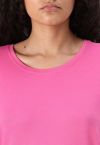 Růžová košile s krátkým rukávem a kulatým výstřihem. Materiál vypadá hladce a má mírnou elasticitu. Bez viditelných vzorů nebo dekorací.