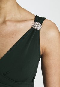 Zelené šaty do obálky s texturovanou tkaninou a klenotem na rameni, zdůrazňující strukturovaný a přiléhavý design.