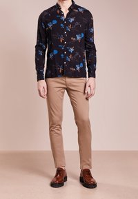 Černá košile s dlouhým rukávem a modrým květinovým vzorem, s límcem na knoflíky. Věnována s khaki slim-fit kalhotami a hnědými šněrovacími botami.