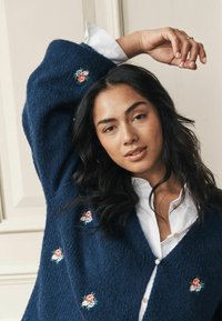 Námornícky pletený kardigán s vyšívanými ružami na predných paneloch, so V-výstrihom a perlovými gombíkmi, nosený cez biele tričko.