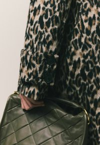 Ruka drži tmavozelenú prešívanú koženú kabelku a má na sebe kabát s leopardím vzorom a čiernymi gombíkmi na rukáve.