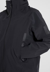 Veste Adidas noire avec capuche, fermeture éclair à l'avant, main dans la poche, avec un logo discret sur la manche gauche.