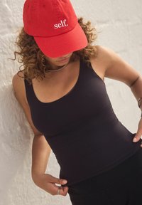Must mustriga tank top, millel on vormiv disain ja tekstureeritud vöökoht, koos punase mütsiga, millel on valge tekst. Taust on hele ja neutraalne.