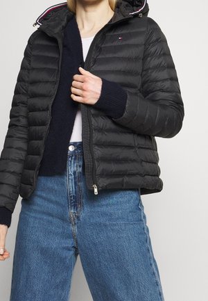 Personne portant une veste noire matelassée à capuche par-dessus un pull bleu marine et un jean bleu clair, tenant la fermeture éclair de la veste près de la poitrine.