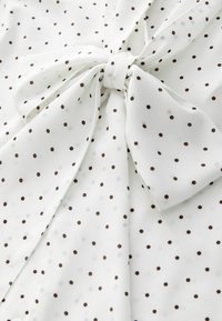 Biała bluzka w brązowe kropki, z wiązaną kokardą przy dekolcie. Miękki, lekki materiał o gładkiej fakturze.