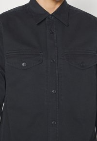 Mann trägt ein schwarzes Hemd mit Knöpfen, zwei Brusttaschen mit Klappen und einem spitzen Kragen.