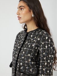 Donna con lunghi capelli scuri che indossa una giacca nera testurizzata decorata con piccole paillettes floreali bianche e orecchini d'argento rotondi, rivolta verso sinistra.