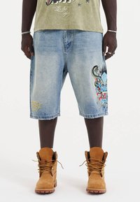 Farkkushortsit vaalean sinisessä pesussa, joissa on suuri värikäs lohikäärmegrafiikka vasemmassa lahkeessa ja hienovarainen keltainen tekstityyli. Ruskeat nauhakenkäbootit.