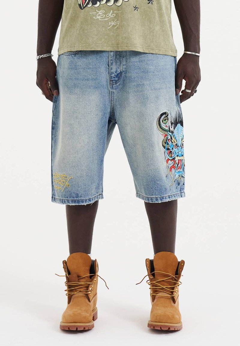 Farkkushortsit vaalean sinisessä pesussa, joissa on suuri värikäs lohikäärmegrafiikka vasemmassa lahkeessa ja hienovarainen keltainen tekstityyli. Ruskeat nauhakenkäbootit.