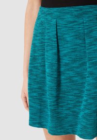 Mujer con una falda texturizada en color azul verdoso con pliegues, mostrando parte de su brazo izquierdo y mano contra un fondo claro y simple.