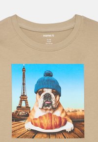 Bulldog a usar um chapéu de malha azul senta-se atrás de um prato com um croissant, Torre Eiffel e o horizonte de Paris ao fundo sobre um tecido bege.