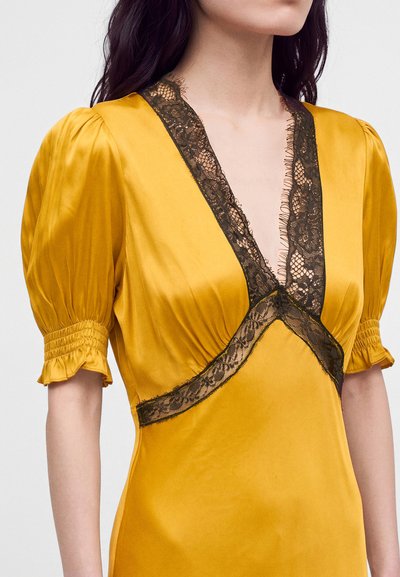 Femme portant une robe en satin jaune moutarde avec des manches bouffantes et un détail en dentelle noire en V.