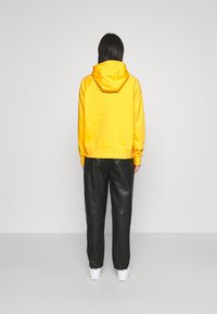 Geel hoodie met capuchon en geribde manchetten, gecombineerd met zwarte leren broek. Witte sneakers zichtbaar bij de voeten. Effen kleur, casual ontwerp.