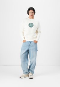 Felpa crema con logo circolare verde, abbinata a jeans baggy azzurri chiari e sneakers beige. Texture in cotone morbido; vestibilità rilassata.