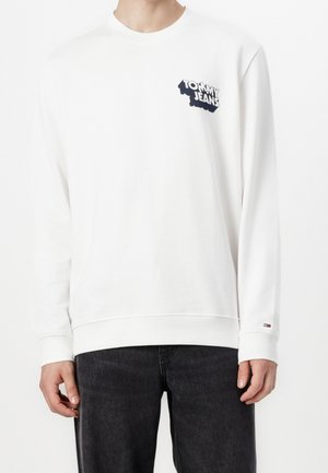 Hvid langærmet sweatshirt med "Tommy Jeans" logo på brystet, båret med sorte jeans, der viser overkroppen og hænderne.
