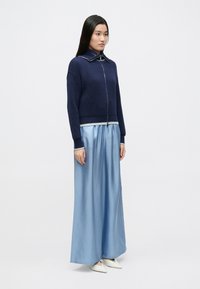 Tumesinine soonikkoega lukuga kampsun valge äärisega, kantud koos pika hele sinise satiinse seelikuga ja valgete teravate ninaga kingadega.