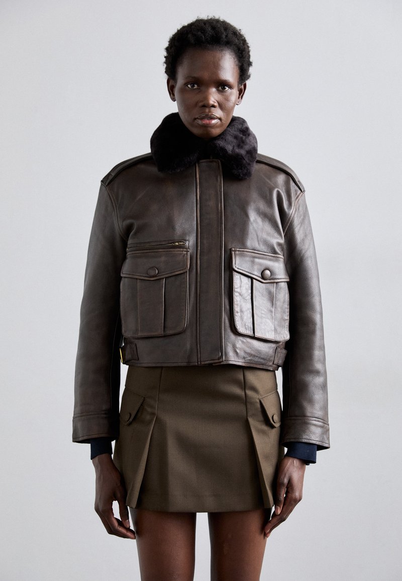 Giacca bomber in pelle marrone con colletto in pelliccia nera, dotata di tasche sul petto e di un design corto, abbinata a una gonna plissettata verde oliva.