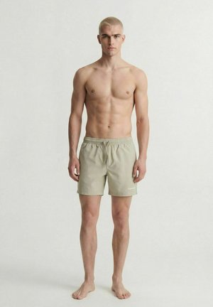 Jeune homme aux cheveux blonds courts, pieds nus, portant un short de bain beige clair, sur un fond blanc uni.