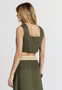 Donna con capelli biondi che indossa un top a maniche corte color verde oliva e una gonna alta coordinata, con una cintura di stoffa beige, girata di spalle.