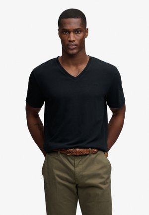 Homme portant un T-shirt noir à col en V, ceinture tressée marron et pantalon vert olive, debout les mains dans le dos.